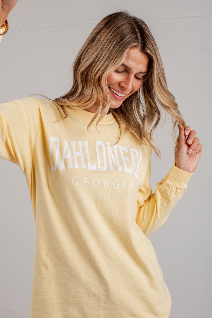 A woman wearing a pale yellow long-sleeve t-shirt with 'DAHLONEGA GEORGIA' printed in white across the chest, posing against a light gray wall.