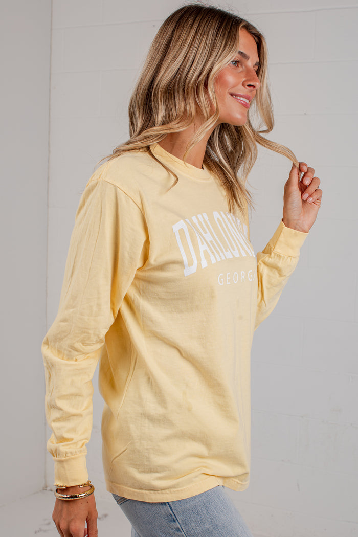 A woman wearing a pale yellow long-sleeve t-shirt with 'DAH GEORGIA' printed in white arched text on the chest, paired with light blue jeans, standing against a white paneled wall.