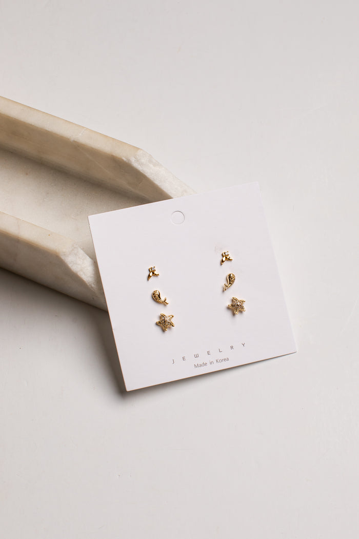 A pair of small gold stud earrings displayed on a white jewelry card, photographed on a light neutral background with a beige tray prop.